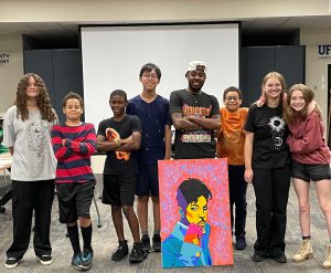 Photo of artist surrounded by art students with a large canvas in the middle. 