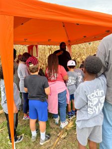 A group of children wearing casual clothing and sneakers are gathered under an orange canopy tent, attentively listening to an instructor speaking. The tent is set up amidst tall, dried cornstalks that stretch into the background, suggesting an agricultural learning environment.