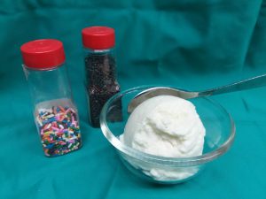 Scoop of vanilla ice cream in small clear bowl with spoon and two containers of sprinkles