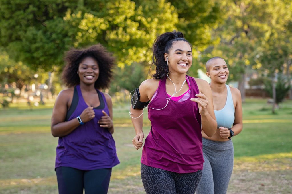 Group of curvy girls friends jogging together at park. Beautiful smiling young women running at the park on a sunny day. Female runners listening to music while jogging.
