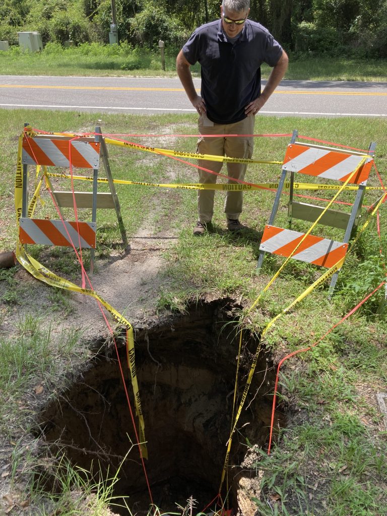 Suwannee River Water Management District staff member is standing next to a sinkhole in Union County Florida. The sinkhole is next to a paved road, and is marked off with 2 barriers and caution tape so that no one gets too close to it.
