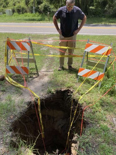 This image shows a sinkhole that formed in Union County FL, near the side of a rural highway. Caution flagging and barricades are set up in front of the sinkhole.