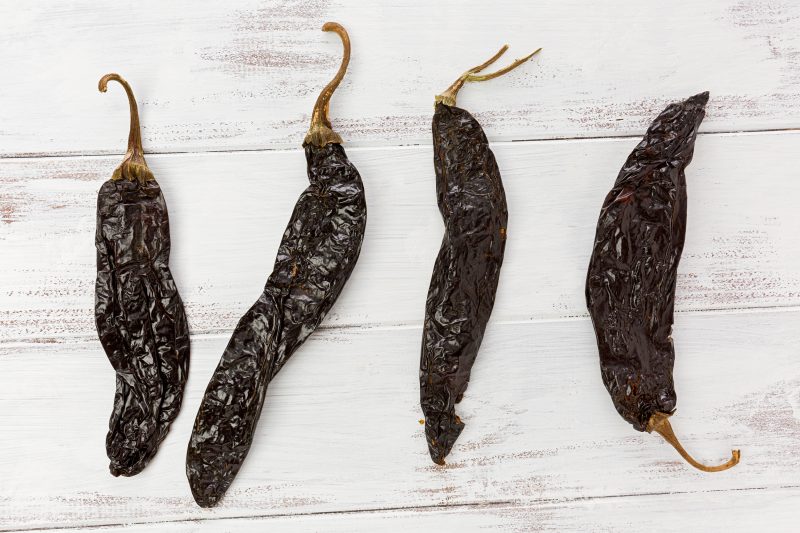 Four dried pasilla peppers with dark, wrinkled skin and curved stems, displayed on a white wooden surface.