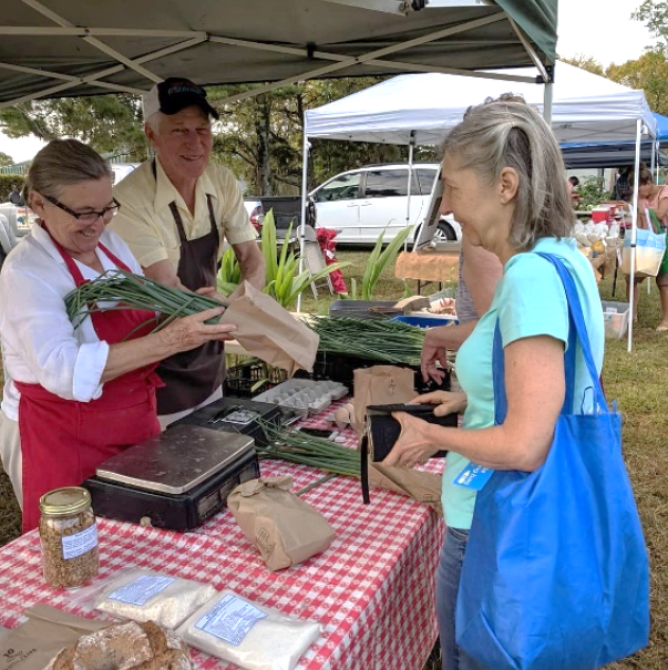 Vendors handing fresh produce to a smiling customer at an outdoor farmers market stall with a red checkered tablecloth.